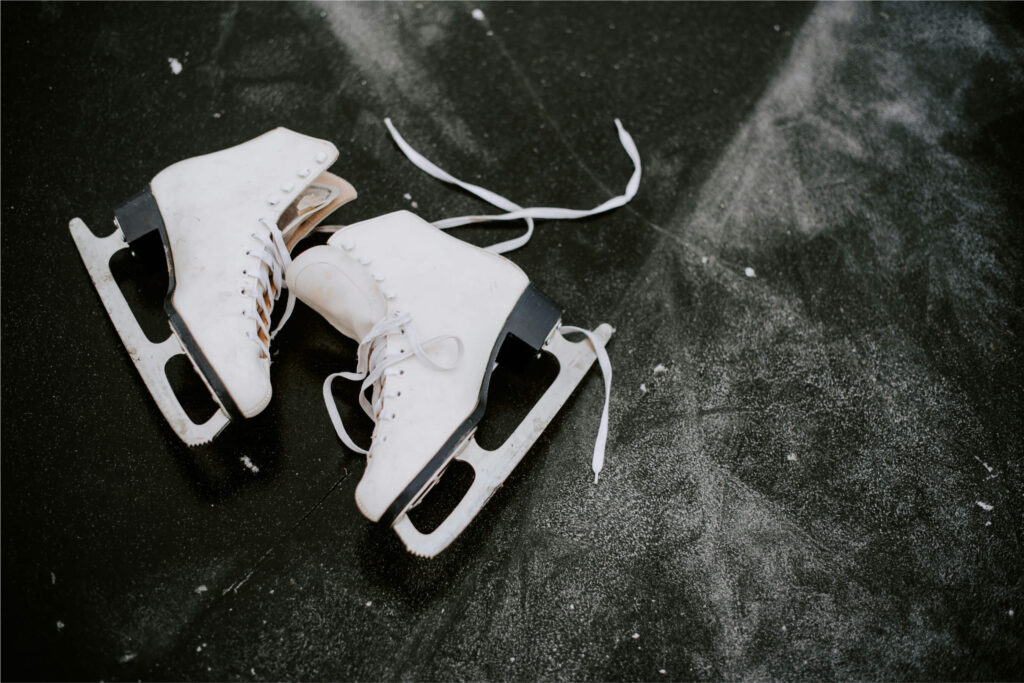 Pair of white figure skates with untied laces on a dark, scuffed ice rink surface.