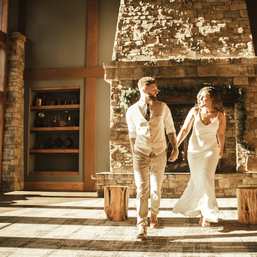 A couple holding hands strolls indoors near a stone fireplace, bathed in warm sunlight. The man wears a vest and tie, while the woman wears a long white dress, reminiscent of Suncadia homes’ elegance, where timeless charm meets modern comfort.