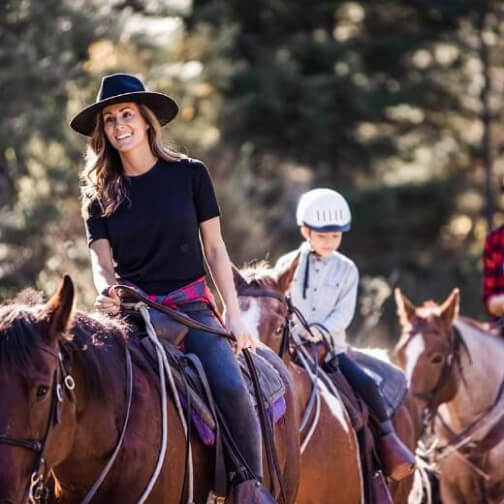 A woman and a child, both wearing helmets, ride horses on a sunny day at Suncadia Resort. The scenic backdrop of trees adds to the charm of this Washington retreat.