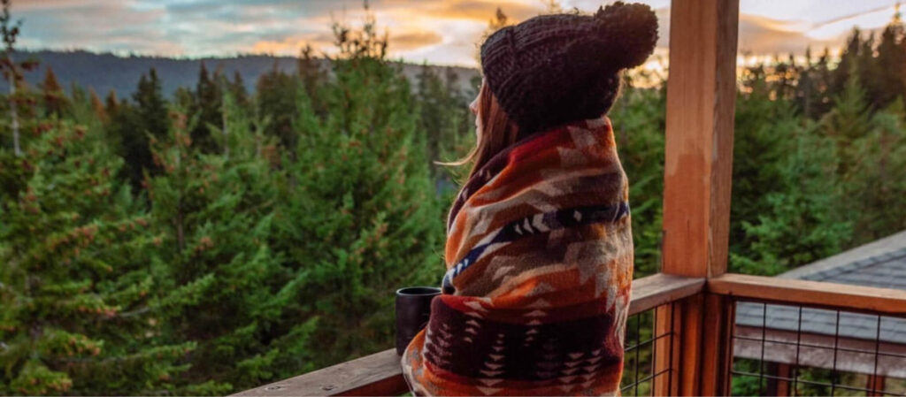 A person wrapped in a colorful blanket and wearing a beanie stands on a wooden deck at Suncadia Resort, holding a mug, overlooking the stunning forest landscape at sunset.