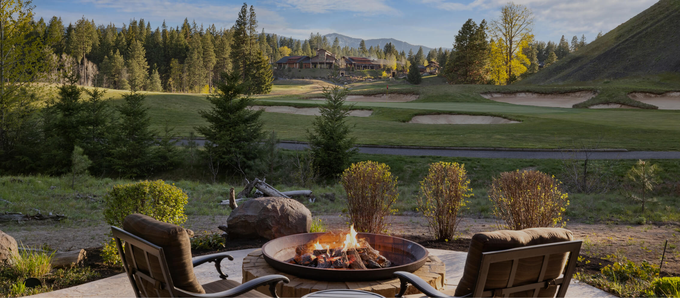 Two chairs face a lit fire pit on a patio overlooking a golf course, surrounded by trees and the charming Suncadia Resort in Washington, with a large cabin in the background.