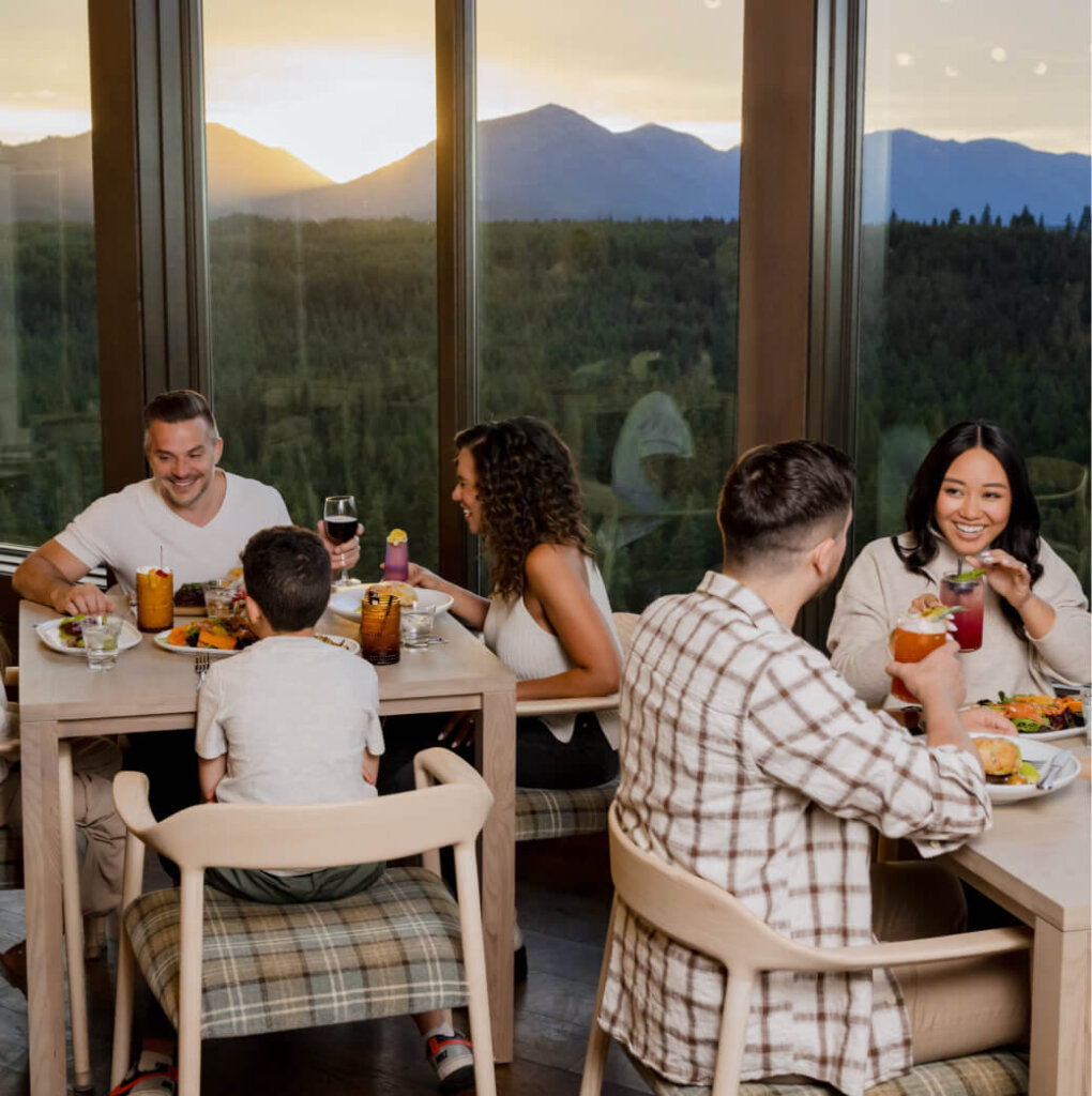 A group of people dines at two tables by a large window with a captivating view of the mountains and forest, reminiscent of Suncadia Resort in Washington. They enjoy various drinks and meals as the sunset casts a golden glow over their evening.