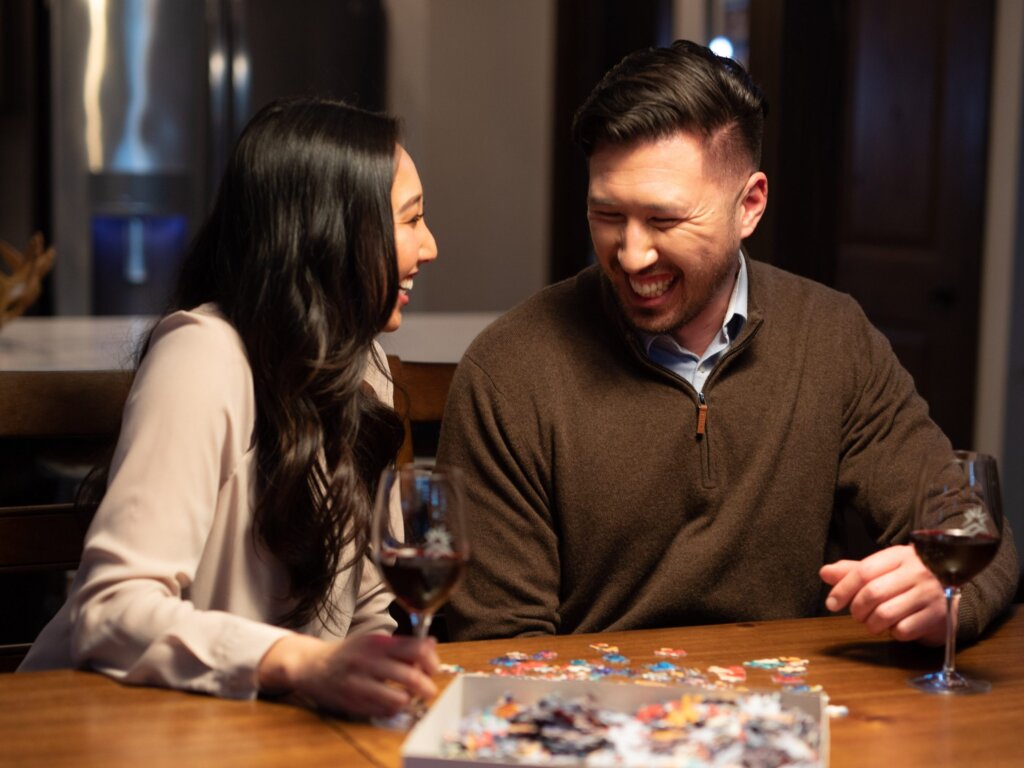 A man and woman sit at a table at Suncadia Resort, laughing together while assembling a jigsaw puzzle. Two glasses of red wine are on the table, complementing the warmth and tranquility of their getaway.
