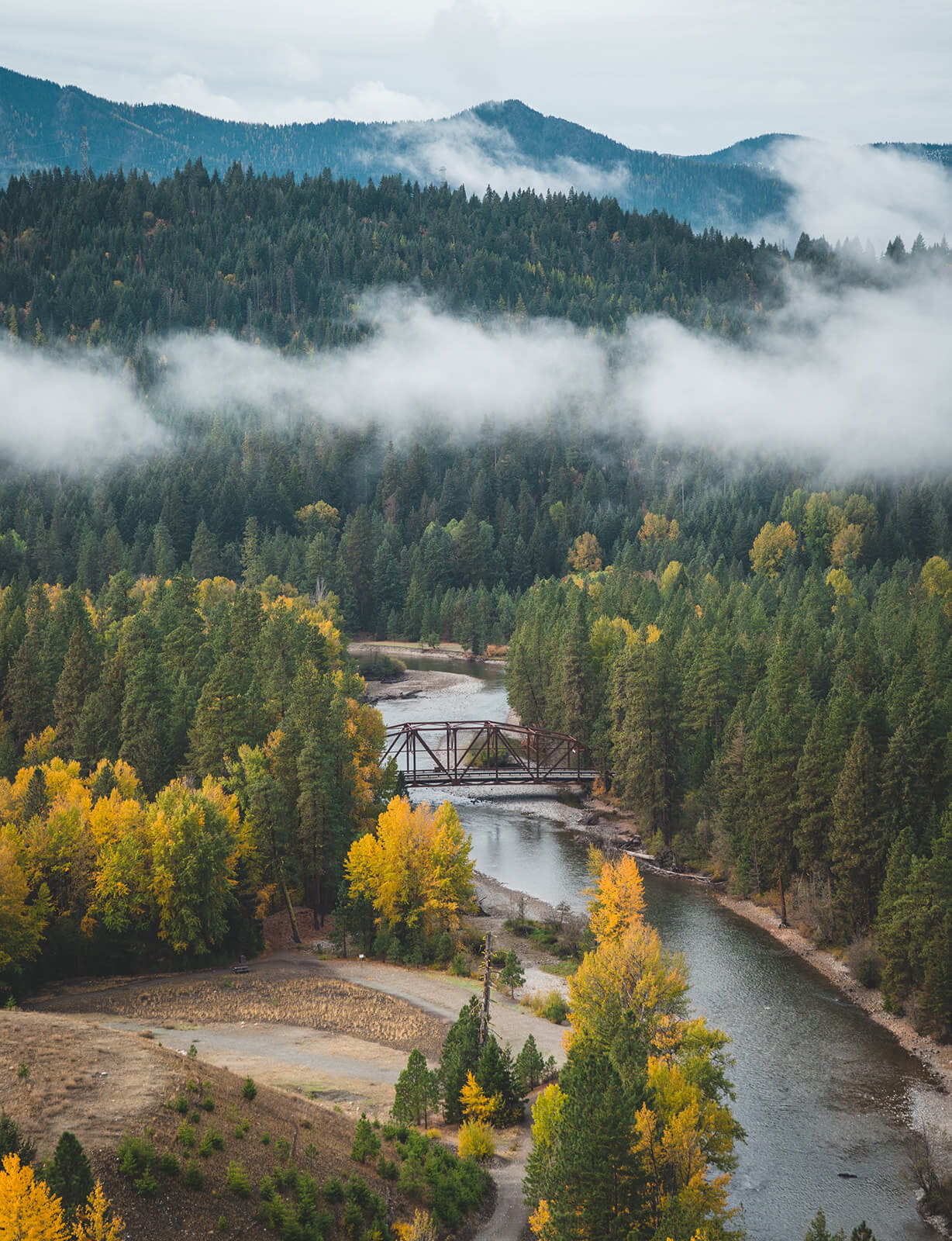 A river flows through a forested valley with autumn-colored trees, a bridge crossing over it. Mist hovers above the landscape, with Suncadia homes nestled nearby. Distant mountains are visible under an overcast sky, adding to the serene beauty of Suncadia Resort.