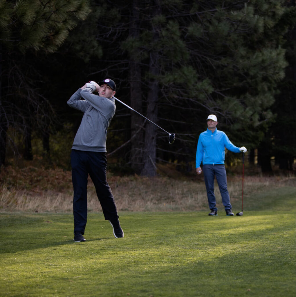 Two people enjoying a round of golf on a lush, tree-lined course at Suncadia Resort, with one swinging a club while the other watches in the background.