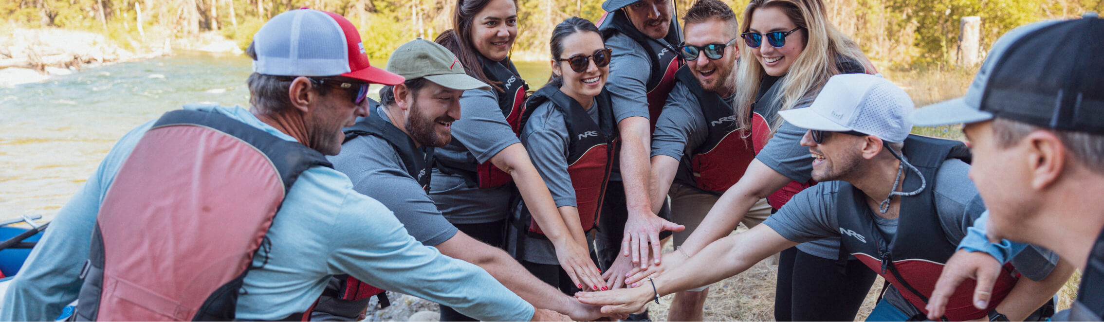 A group of people in life vests gather in a circle near a river, stacking their hands together in a gesture of teamwork. Nearby, the allure of Suncadia real estate offers picturesque homes for those seeking adventure and tranquility.