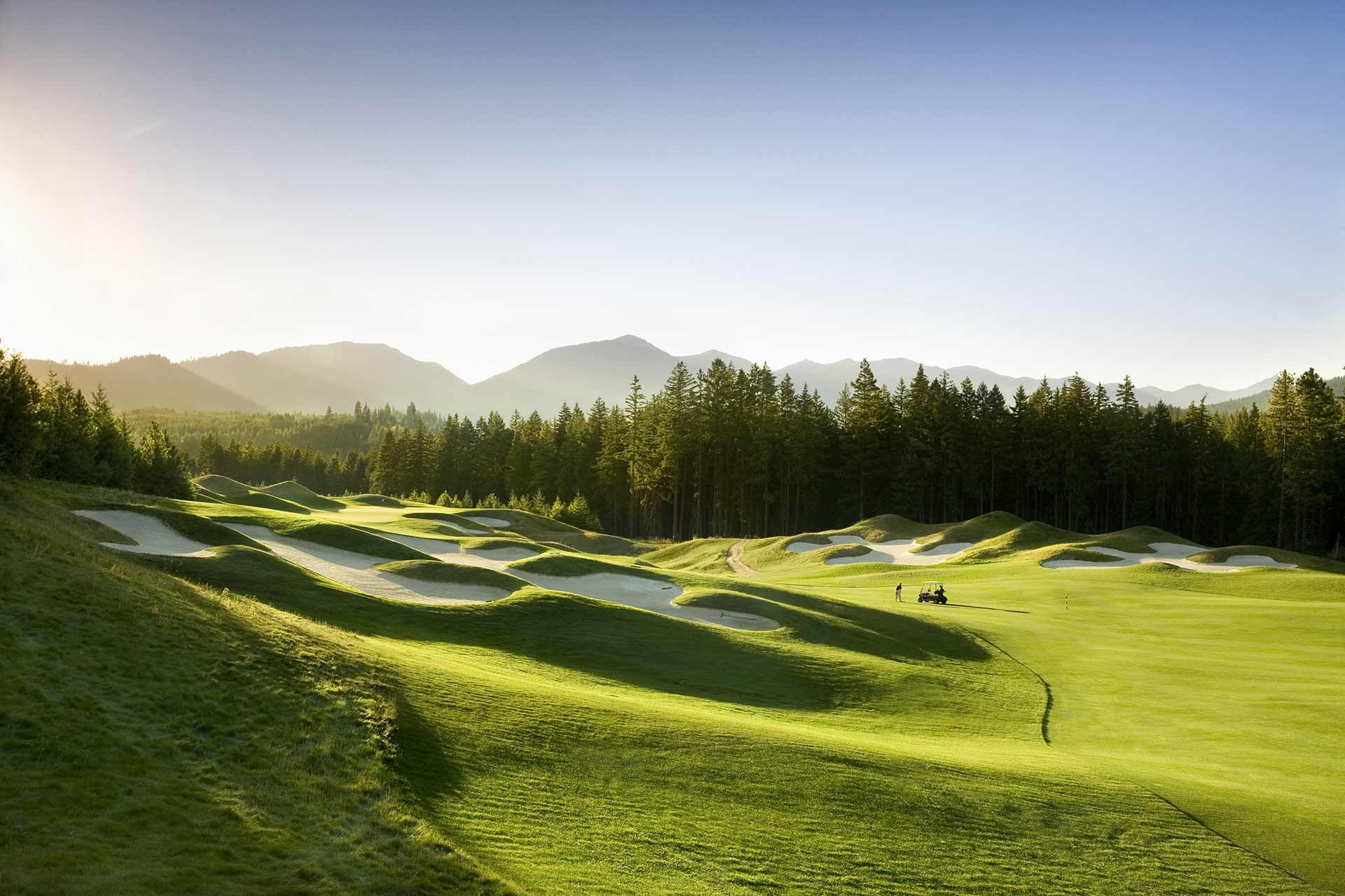 Expansive golf course with rolling hills, sand traps, and distant trees under a clear blue sky at Suncadia Resort. The mountains in the background are bathed in the warm hues of sunset.