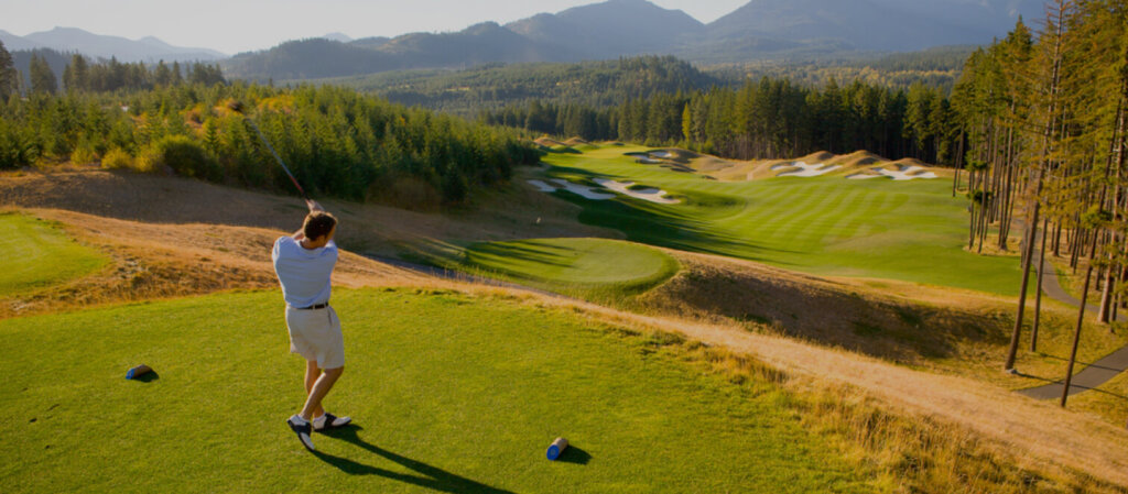 A person in a white shirt and shorts plays golf on a scenic course at Suncadia, with majestic mountains and lush trees providing the perfect backdrop. After the game, they plan to explore Cle Elum restaurants for a delightful dining experience.