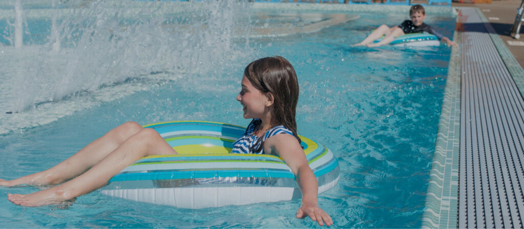 At Suncadia Resort, two children in swimsuits float joyfully on inflatable rings in the pool, with water splashing playfully around them.