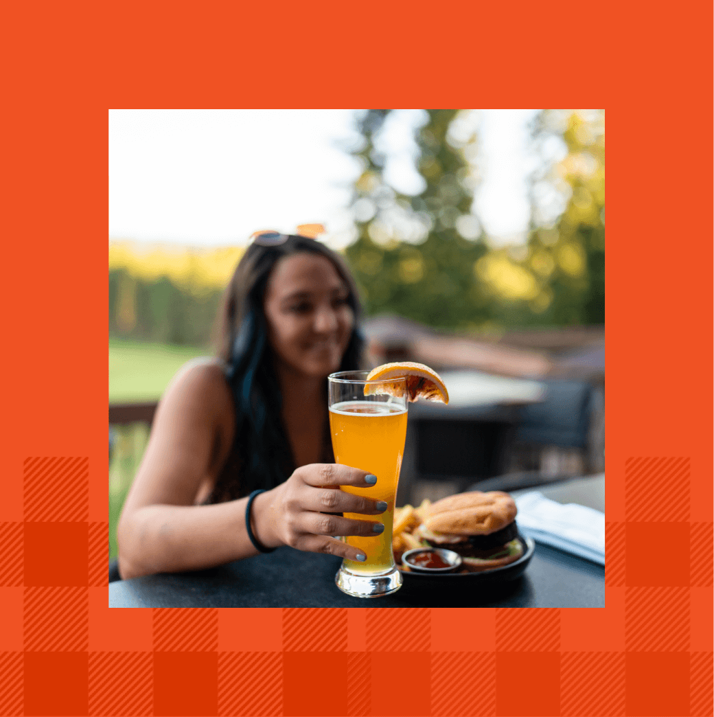 A woman sits at a table, savoring an orange drink adorned with a fruit slice, while beside her lies a tempting plate of food. The image, framed by an orange checkered border, hints at the sunny leisure you can enjoy at Suncadia Resort in Washington.