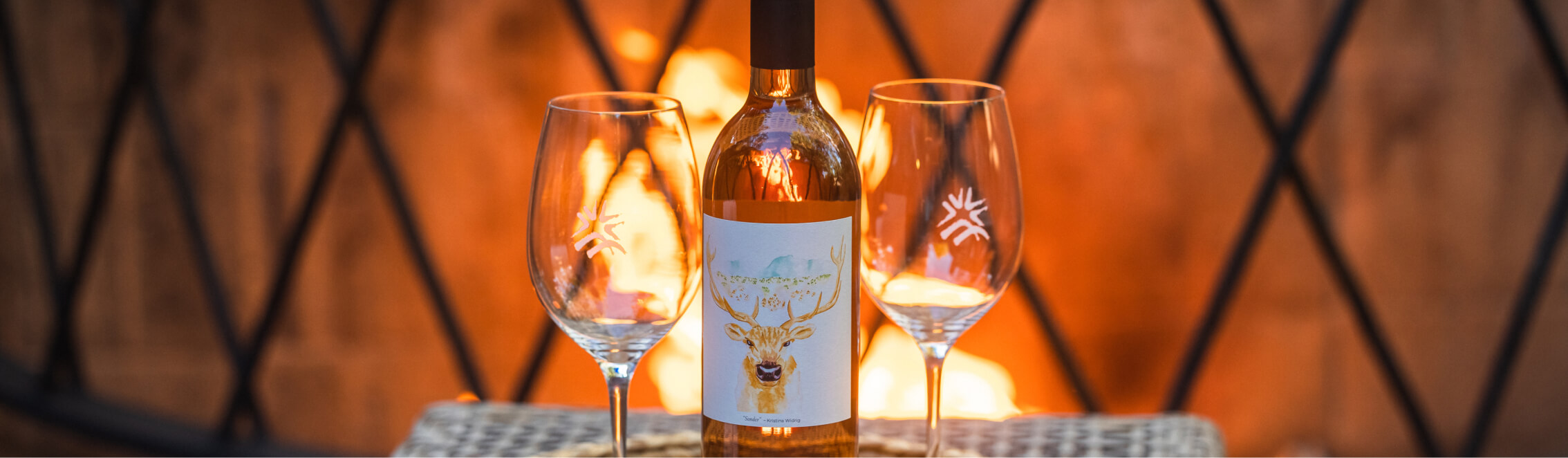 A bottle of wine with a deer label stands between two empty wine glasses, set against a warm, softly lit background reminiscent of evenings at Suncadia Resort.