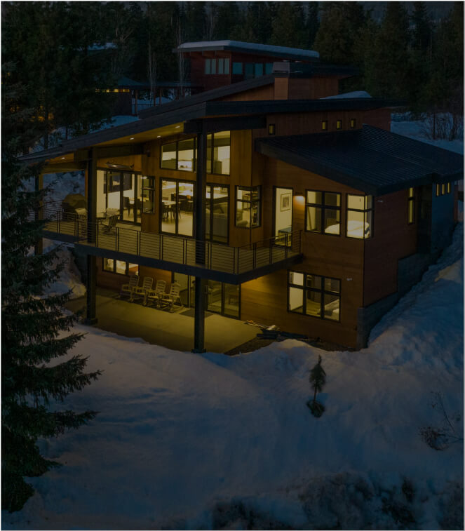 A modern, two-story house with large windows is illuminated at night, surrounded by snow and trees, echoing the serene charm of Suncadia Resort.