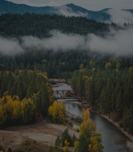 A misty, forested landscape at Suncadia Resort features a river winding through the trees, with a charming bridge crossing over. Majestic mountains stand in the background, painting an idyllic scene perfect for exploring Suncadia real estate and finding your dream home.