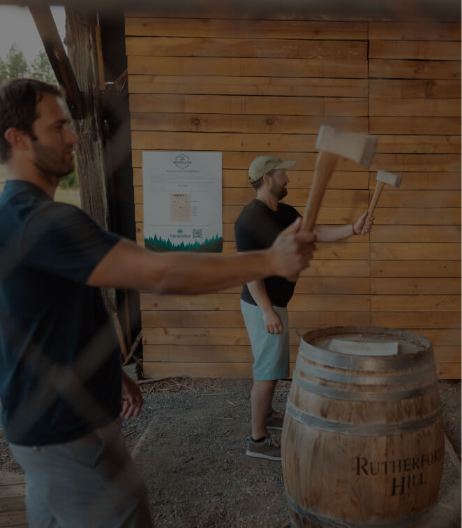 Two men stand next to a wooden barrel, holding axes as if preparing to throw them at a target. The rustic setting, reminiscent of Suncadia Resort's outdoor charm, features a wooden backdrop that perfectly complements the adventurous spirit of Cle Elum.