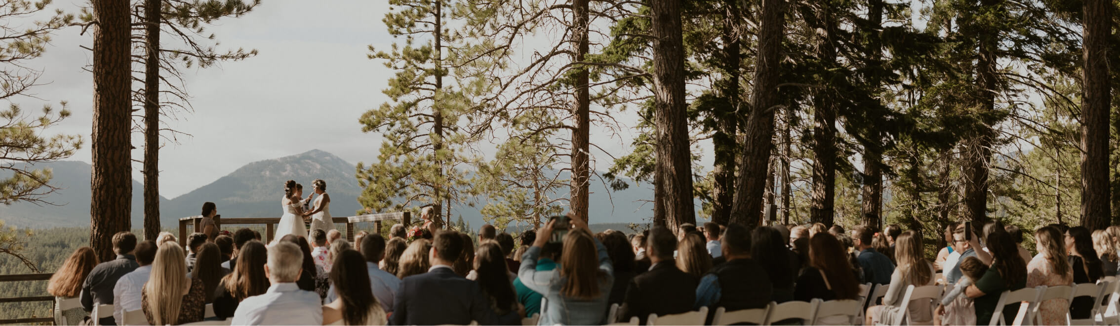 At a scenic outdoor altar at Suncadia Resort Washington, a couple exchanges vows, surrounded by lush forest and seated guests. The majestic mountains rise in the background under a clear sky, completing this breathtaking scene near the inviting Cle Elum restaurants.