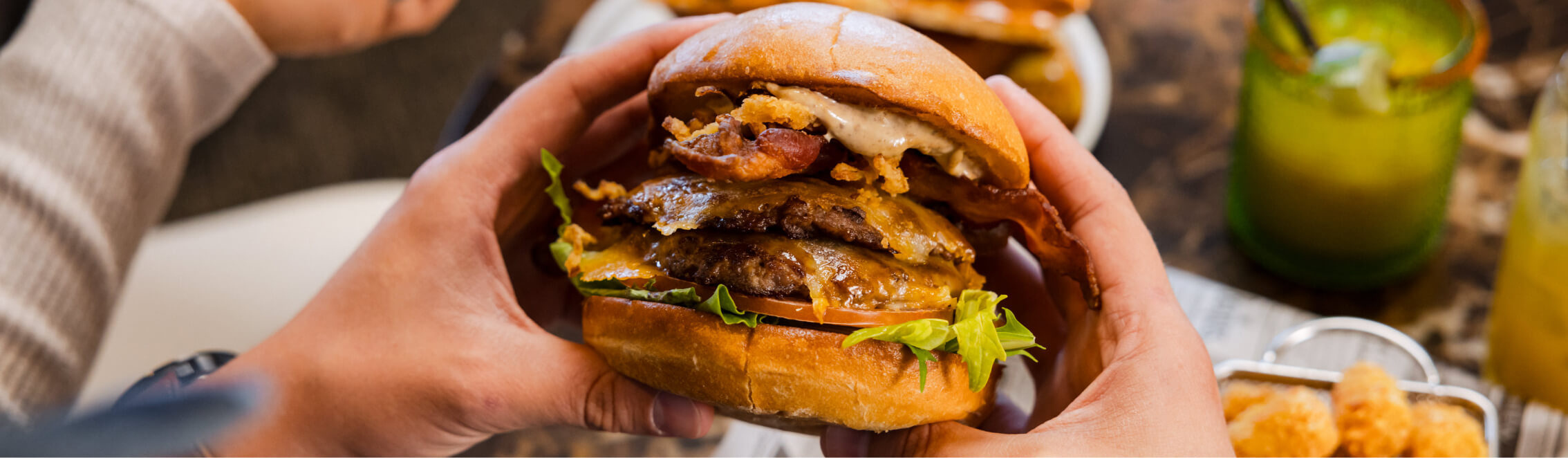 A person enjoys a juicy cheeseburger with bacon, lettuce, and onion at one of the charming Cle Elum restaurants. In the background, a drink complements the meal perfectly, echoing the delightful ambiance you’d find at Suncadia Resort.