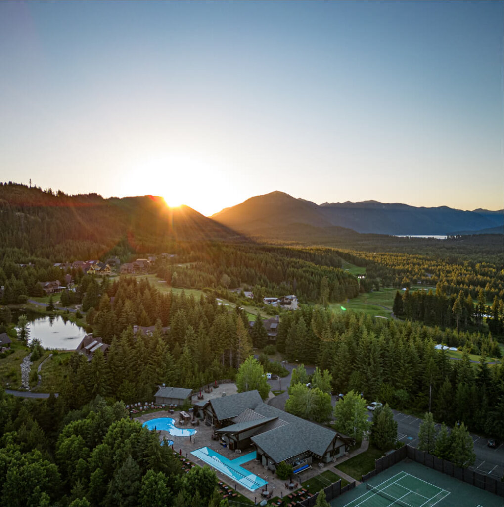 Aerial view of Suncadia Resort at sunset reveals a stunning landscape with a pool, tennis court, and the majestic embrace of surrounding forests and mountains.