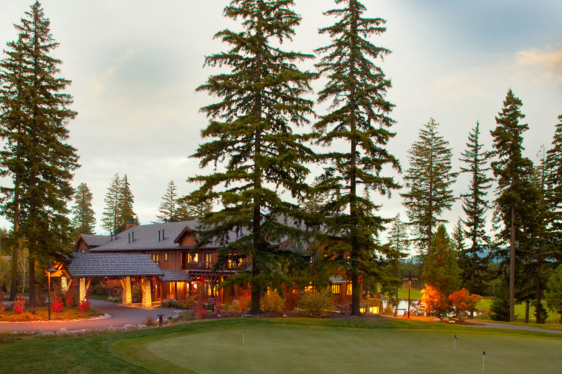 A large rustic lodge surrounded by tall pine trees and a manicured green lawn basks in the golden glow of sunset, epitomizing the serene beauty of Suncadia real estate.