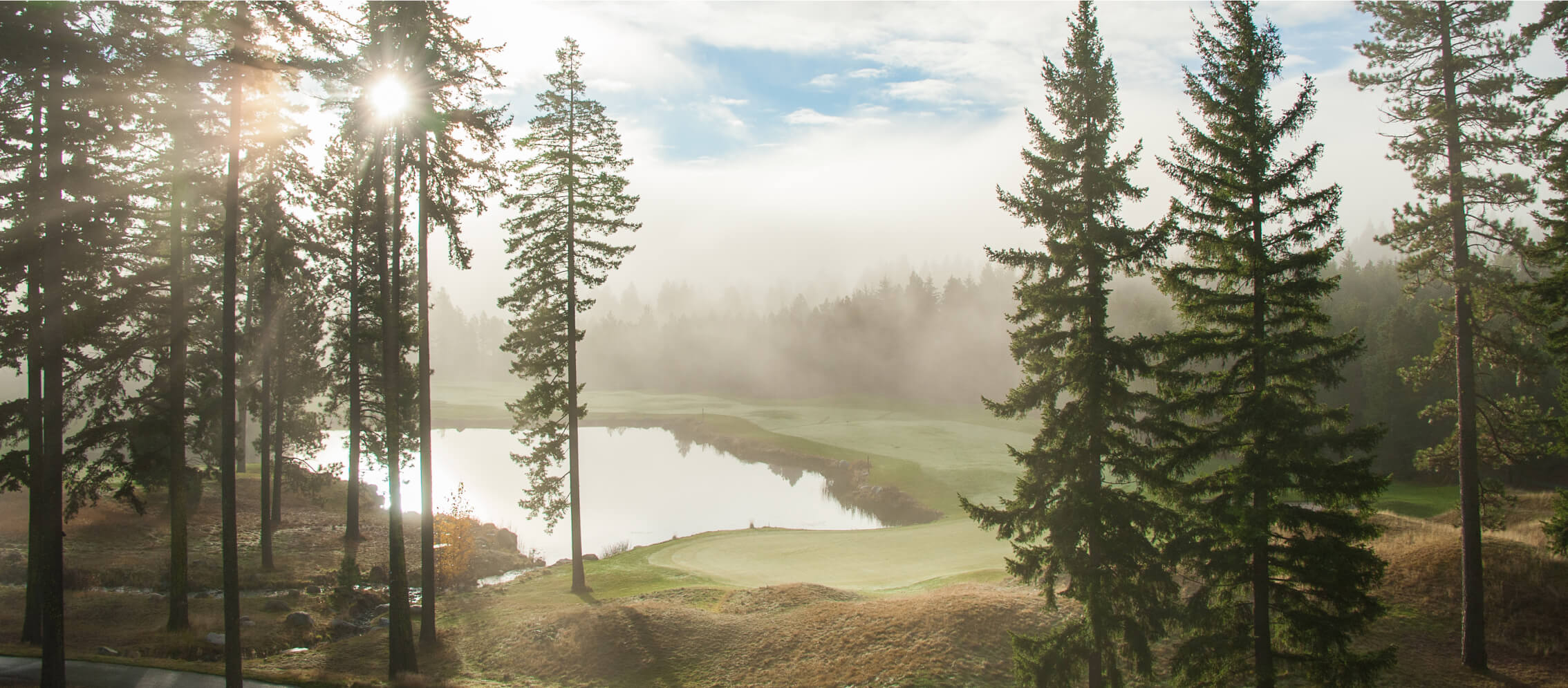 A misty morning landscape with tall pine trees enveloping a tranquil pond under a partly cloudy sky at Suncadia Resort, Washington. Sunlight gently filters through the trees, enhancing the serene beauty of Suncadia homes nestled nearby.