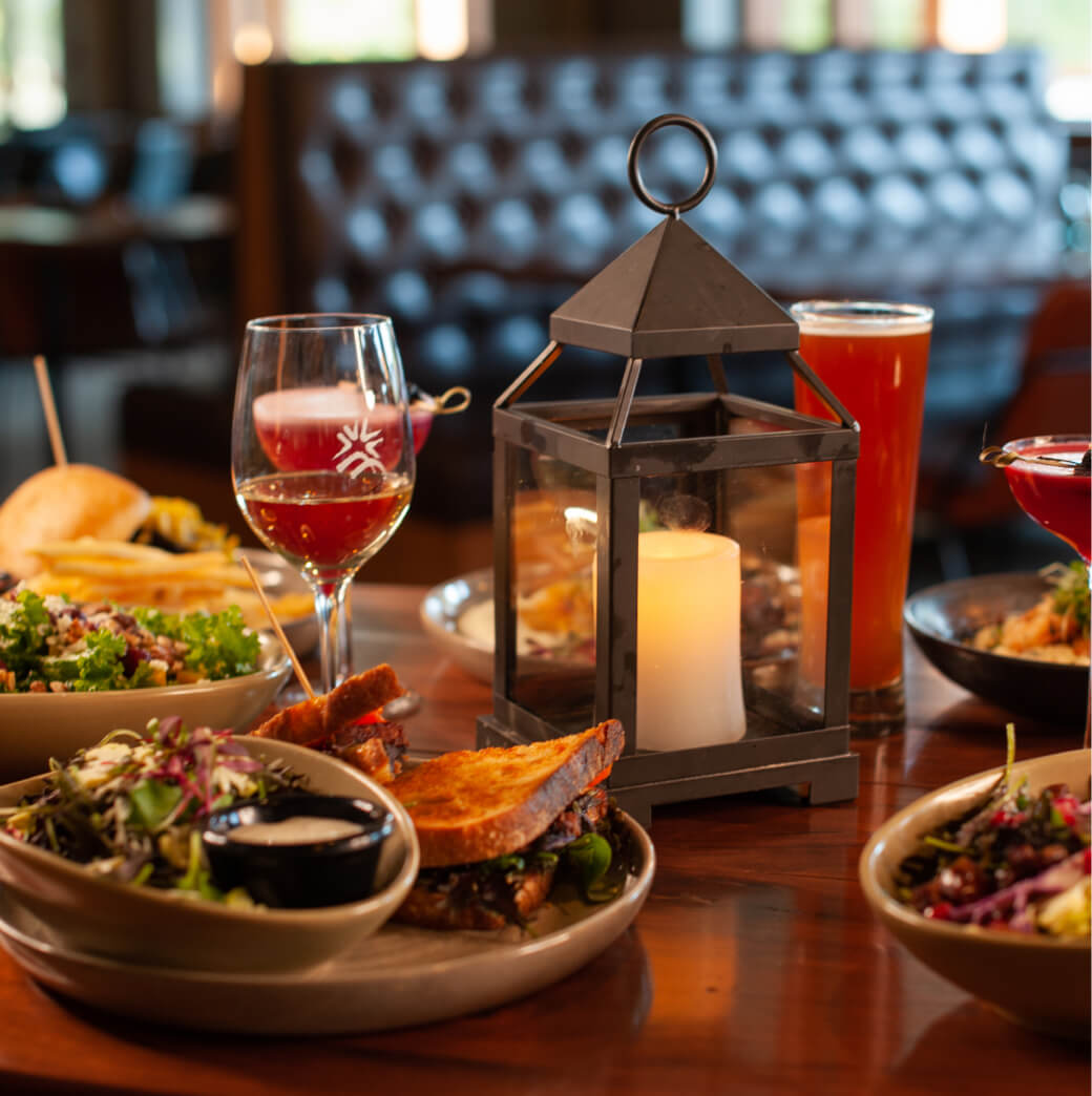 A cozy restaurant table at Suncadia Resort features a lantern casting a warm glow, plates of gourmet food, and several glasses of drinks, perfect for enjoying the serene beauty synonymous with Suncadia.