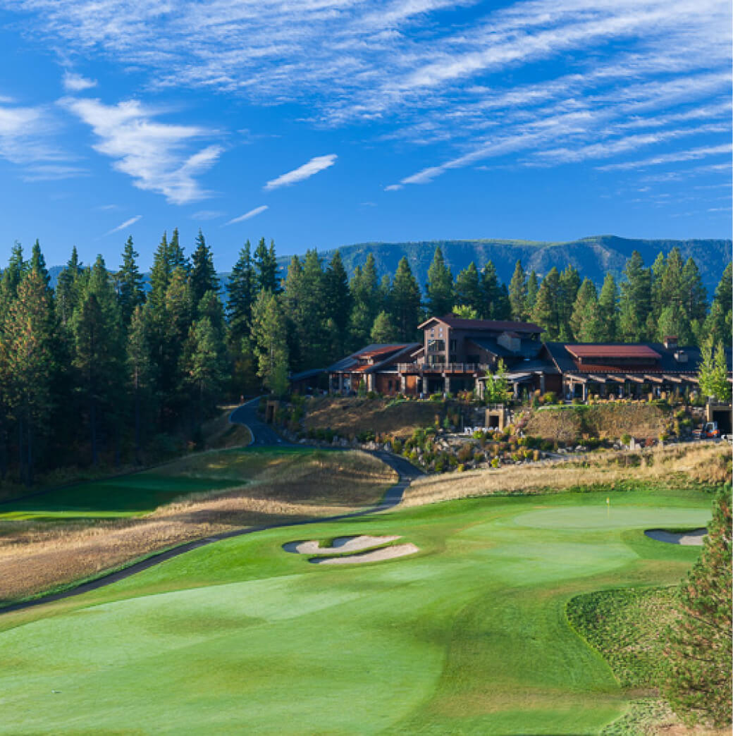 A scenic view of Suncadia Resort's golf course, with a clubhouse in the background, surrounded by pine trees and mountains under a blue, cloud-streaked sky.