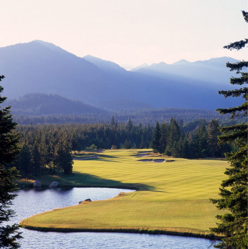 A scenic golf course at Suncadia Resort is surrounded by lush trees, with a calm lake in the foreground and majestic mountains in the background under a hazy sky.