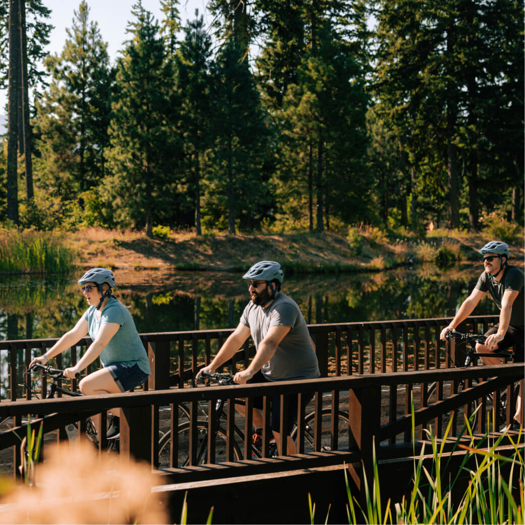 Three people wearing helmets ride bicycles over a wooden bridge in the lush forested area of Suncadia Resort.