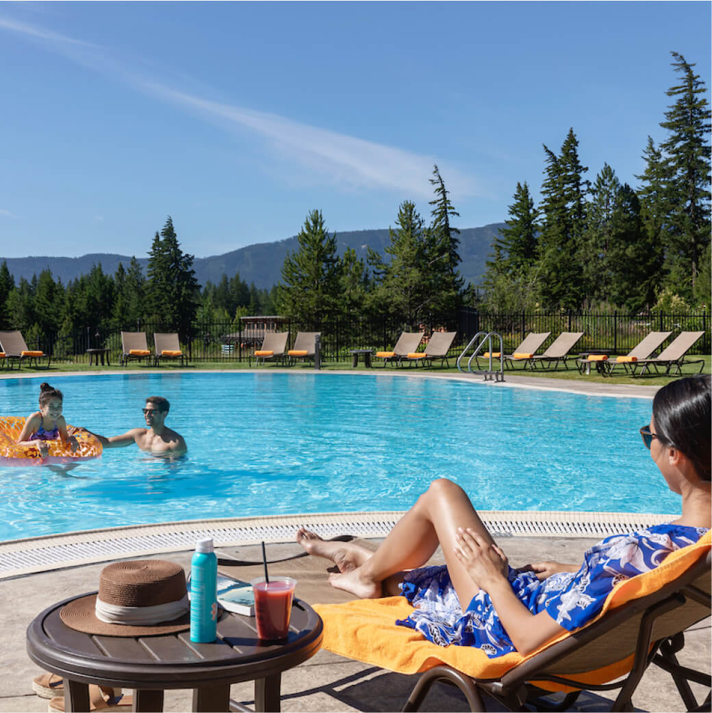 At Suncadia, people relax by a swimming pool surrounded by trees and mountains. A person lounges on a chair with sunblock and a drink nearby, enjoying the serene atmosphere, while others float in the water with an inflatable ring, savoring the perfect day near Cle Elum's attractions.