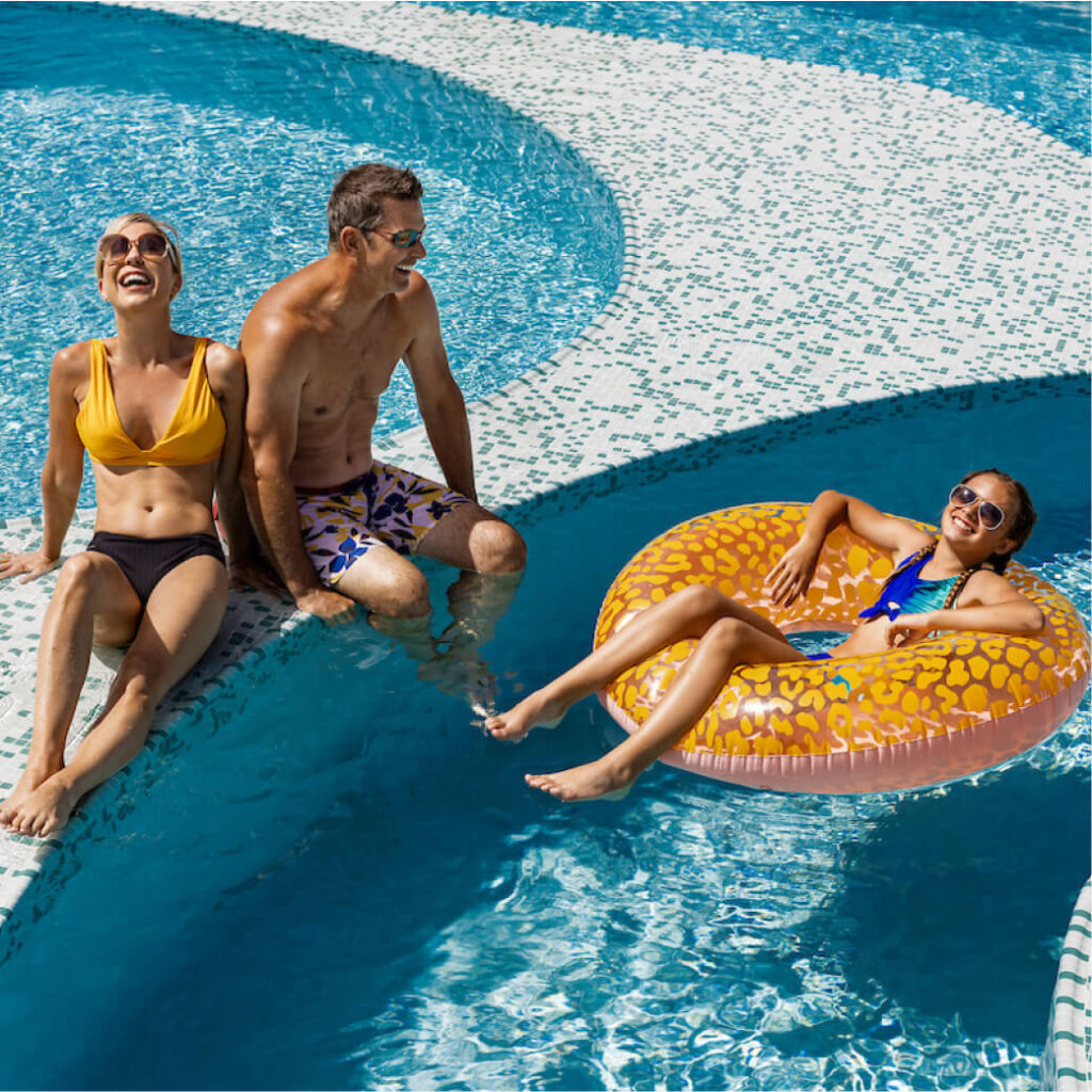 A family enjoys a sunny day at the pool at Suncadia Resort Washington. Two adults sit on the edge with their feet in the water, while a child floats nearby in an inflatable ring, soaking up the tranquil ambiance that Suncadia homes are known for.