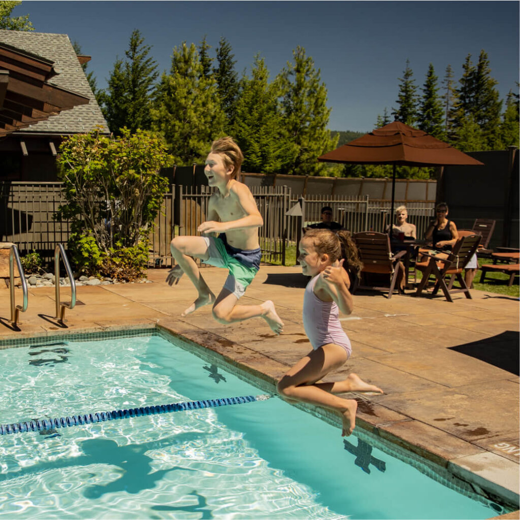 Two children are joyfully jumping into a swimming pool at Suncadia Resort. In the background, people relax under an umbrella, savoring dishes that rival the finest Cle Elum restaurants. The scene is set outdoors on a sunny day, amidst the beautiful landscape of Suncadia homes.