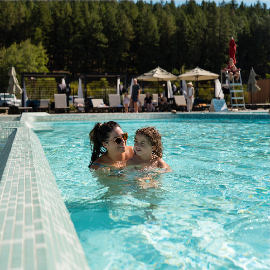 A woman and child enjoy a swim in an outdoor pool at the serene Suncadia Resort. Umbrellas and chairs line the poolside, with lush trees providing a scenic backdrop, making it an idyllic escape for those exploring Suncadia real estate.