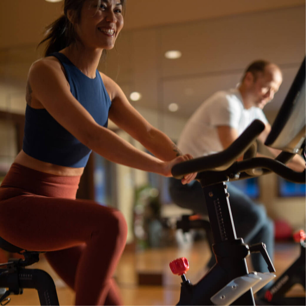 At the Suncadia Resort gym, two people are exercising on stationary bikes. The woman in the foreground is smiling as she enjoys her workout, perfectly capturing the vibrant energy of Suncadia.