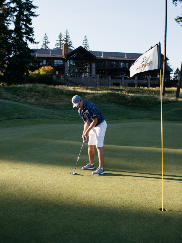 A golfer in a blue shirt and white shorts is putting on a green near a flag, with a large building and trees in the background, showcasing the serene beauty of Suncadia real estate.