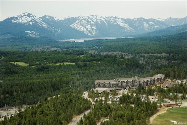 Aerial view of a picturesque lodge nestled amid dense forests and majestic mountains, with snow-capped peaks gracing the horizon, reminiscent of the enchanting Suncadia Resort in Washington.