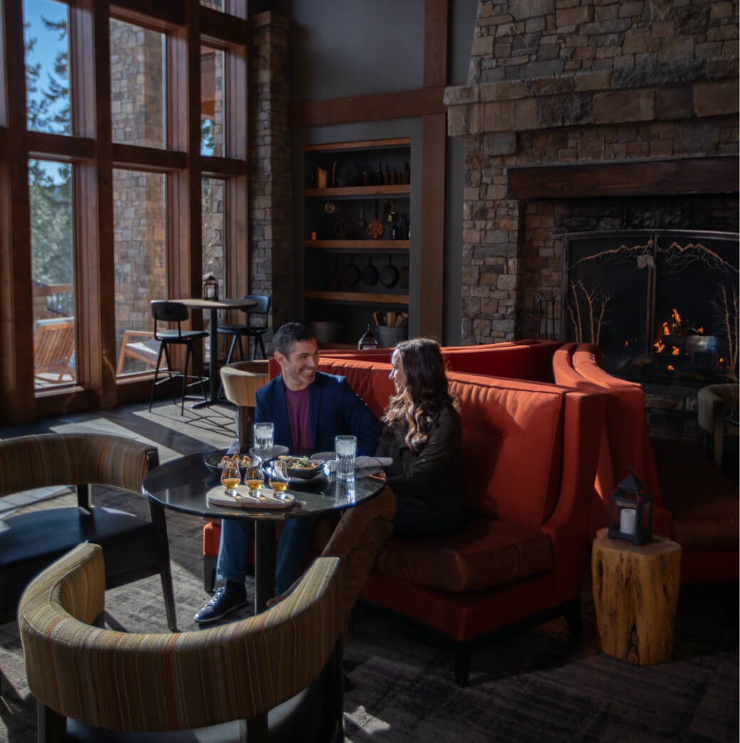 A couple sits at a table in a cozy Cle Elum restaurant with a stone fireplace. They are engaged in conversation and enjoying drinks, savoring the warmth reminiscent of Suncadia homes.