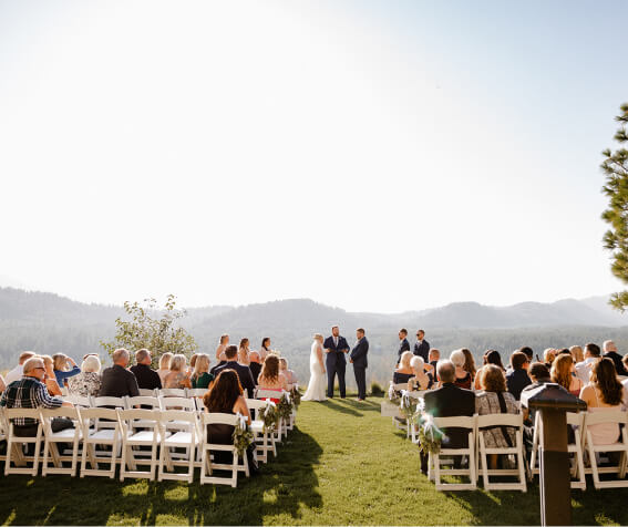 An enchanting outdoor wedding ceremony unfolds at Suncadia Resort in Washington. Guests are seated on white chairs as the couple stands with an officiant and two attendants, all framed by a breathtaking mountain backdrop under a clear sky.