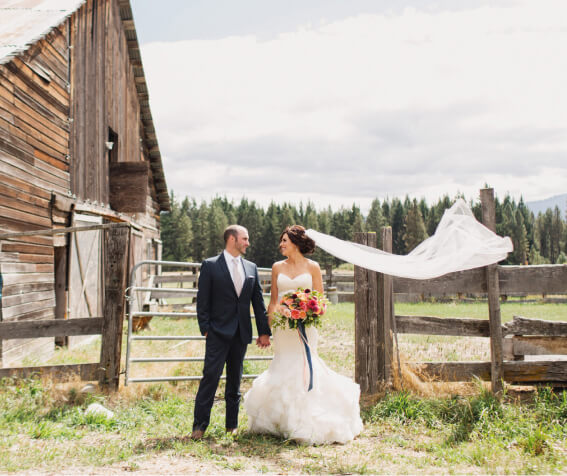 A bride and groom stand holding hands in front of a rustic barn at Suncadia Resort Washington, with the bride's veil flowing in the wind.