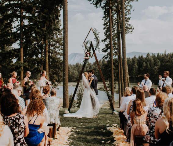 At a romantic outdoor wedding ceremony at Suncadia Resort, a couple shares a kiss beneath a triangular wooden arch adorned with greenery. They're surrounded by guests, set against the stunning backdrop of forest and mountains.