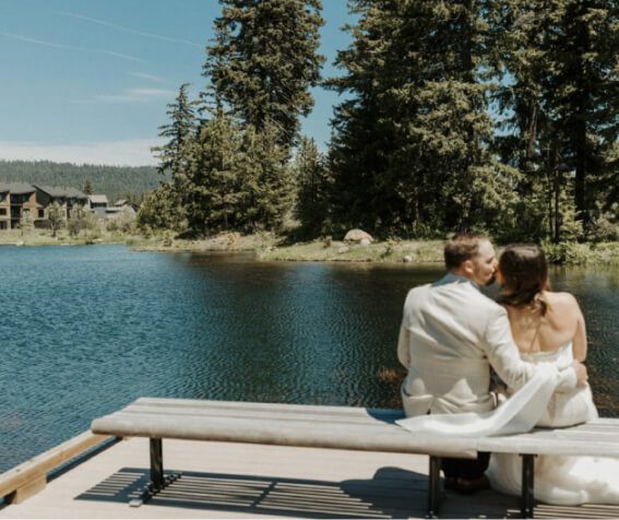 A couple dressed in formal attire sits together on a bench by a lake in Suncadia, surrounded by pine trees and under a clear blue sky, perhaps dreaming of nearby Cle Elum restaurants.