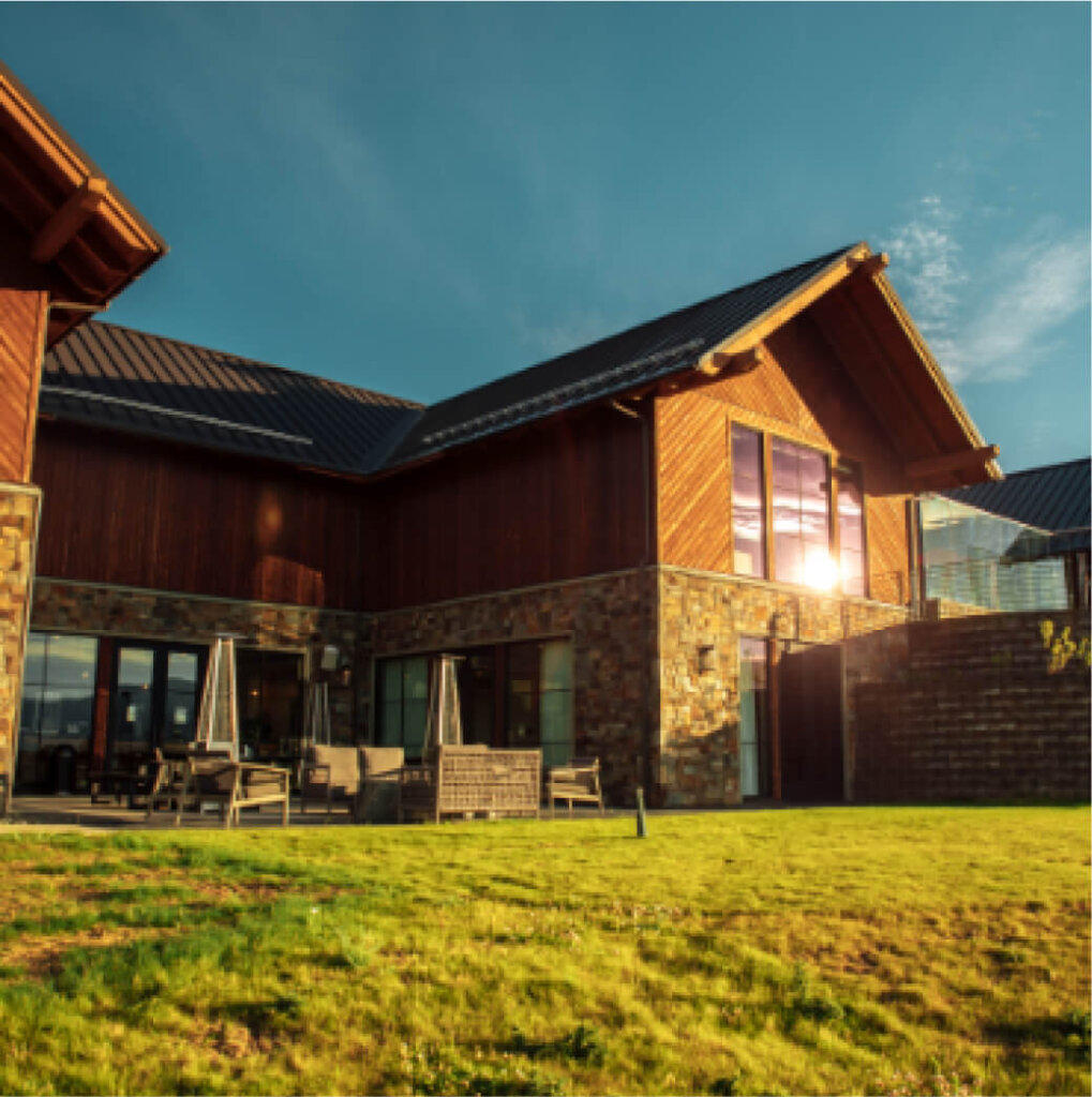 A modern house with large windows and a stone and wood exterior, reminiscent of Suncadia Resort's style. As the sun sets behind, it casts a warm glow on the grassy yard and outdoor seating area, mirroring the serene beauty found near Cle Elum restaurants.