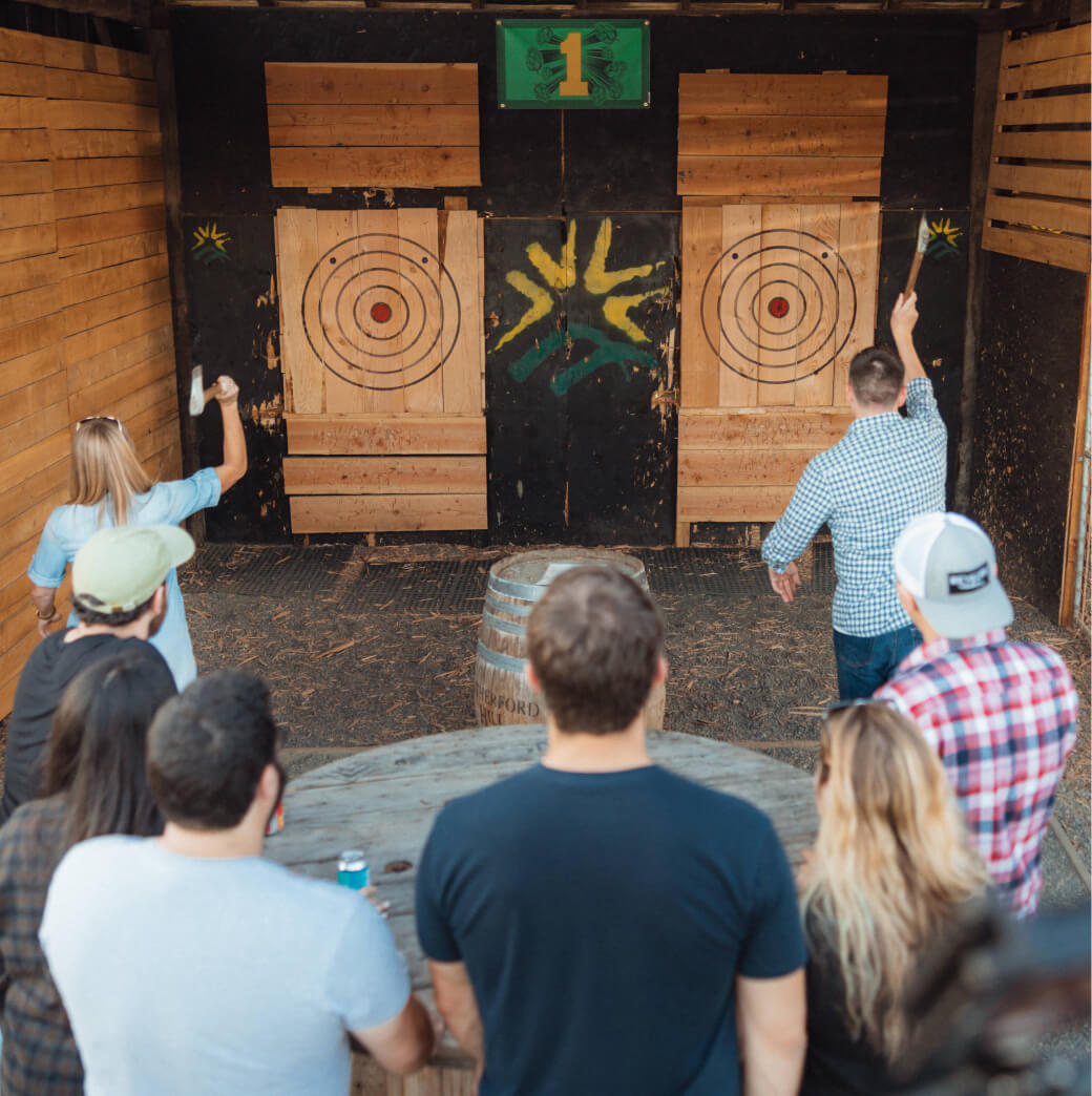 A group of people watch two individuals throwing axes at target boards, marked with the number "1," inside a wooden enclosure at Suncadia Resort in Washington.