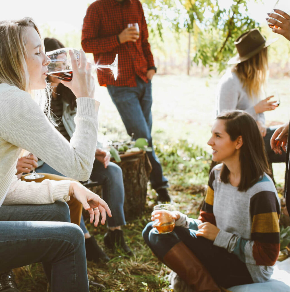 A group of people sits outdoors at Suncadia, socializing and drinking wine. One person sips from a glass, while others hold their drinks. The grassy setting, with trees in the background, adds to the charm of this Suncadia real estate gem.