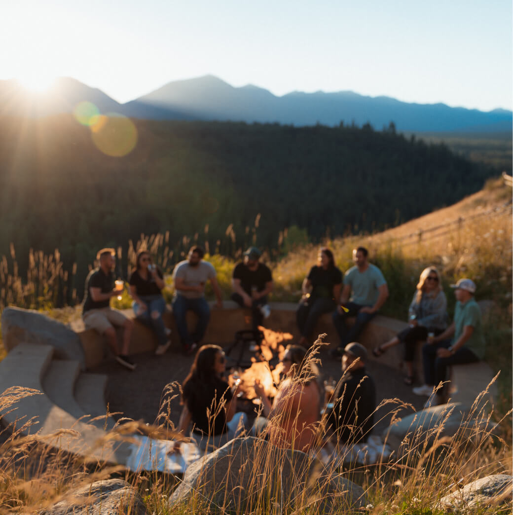 A group of people sits around a campfire on a hillside, with the majestic mountains near Suncadia Resort Washington silhouetted against the sunset, sharing stories and laughter as twilight descends.