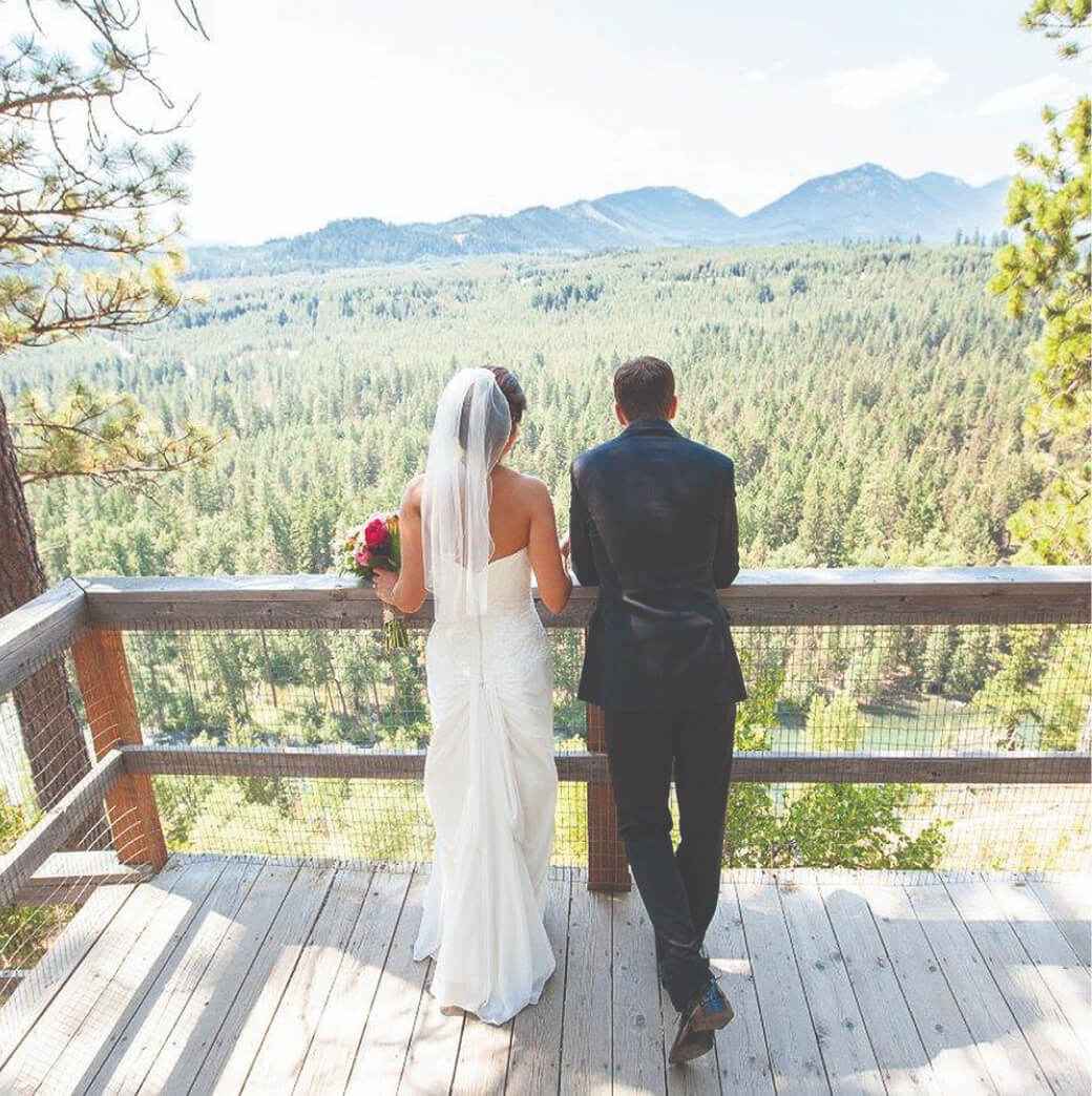 A bride and groom stand on a wooden deck at Suncadia Resort, facing a scenic forest and mountain view in their wedding attire.