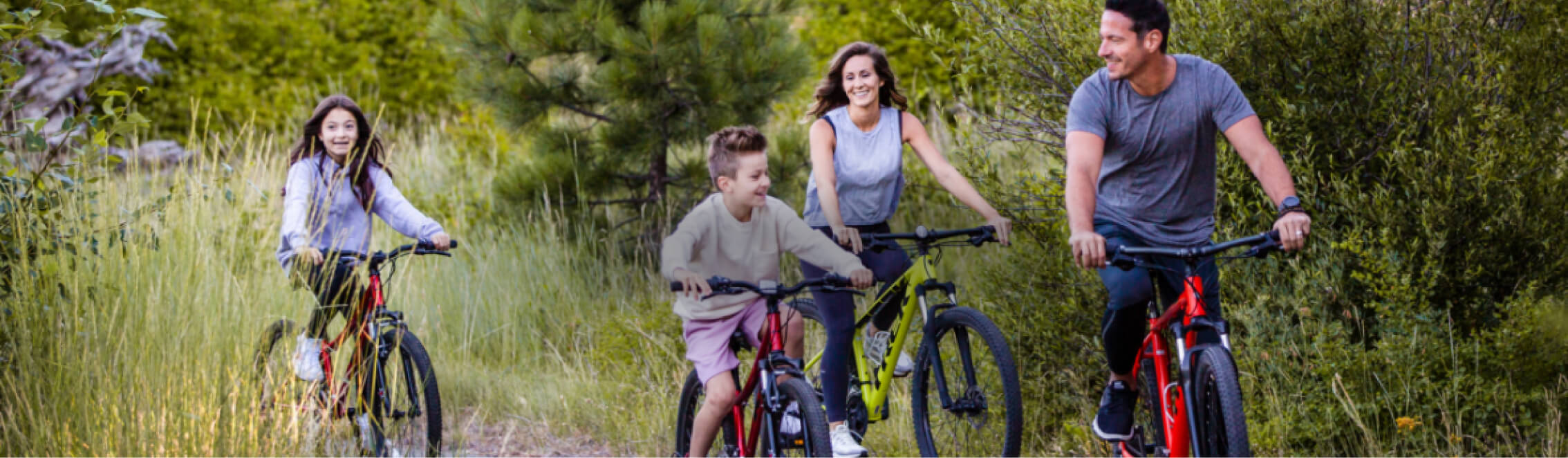 A family of four rides bicycles on a grassy trail at Suncadia Resort, surrounded by lush greenery and trees, smiling and enjoying the outdoors.