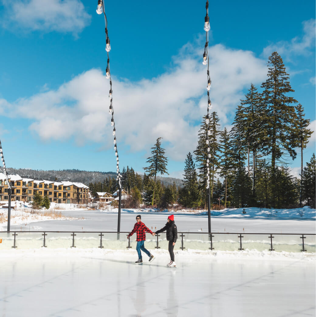 Two people joyfully ice skating on an outdoor rink, surrounded by snow-laden trees under a clear blue sky, create a perfect winter tableau. Nearby, charming buildings reflect the serene lifestyle found in Suncadia homes, where nature and community beautifully intertwine.