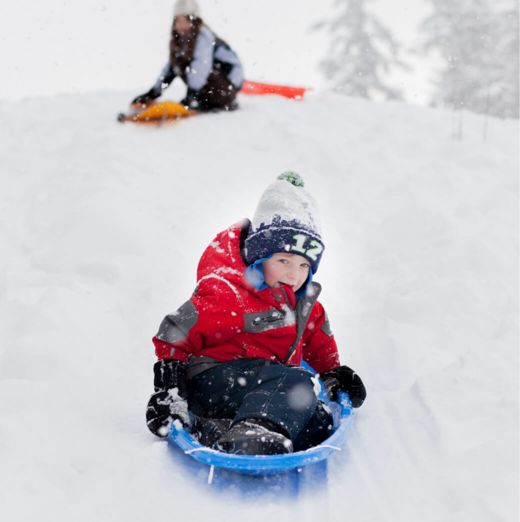 A child in a red jacket and beanie joyfully sleds down a snowy hill at Suncadia Resort Washington, while another person and a sled are visible in the blurred background.