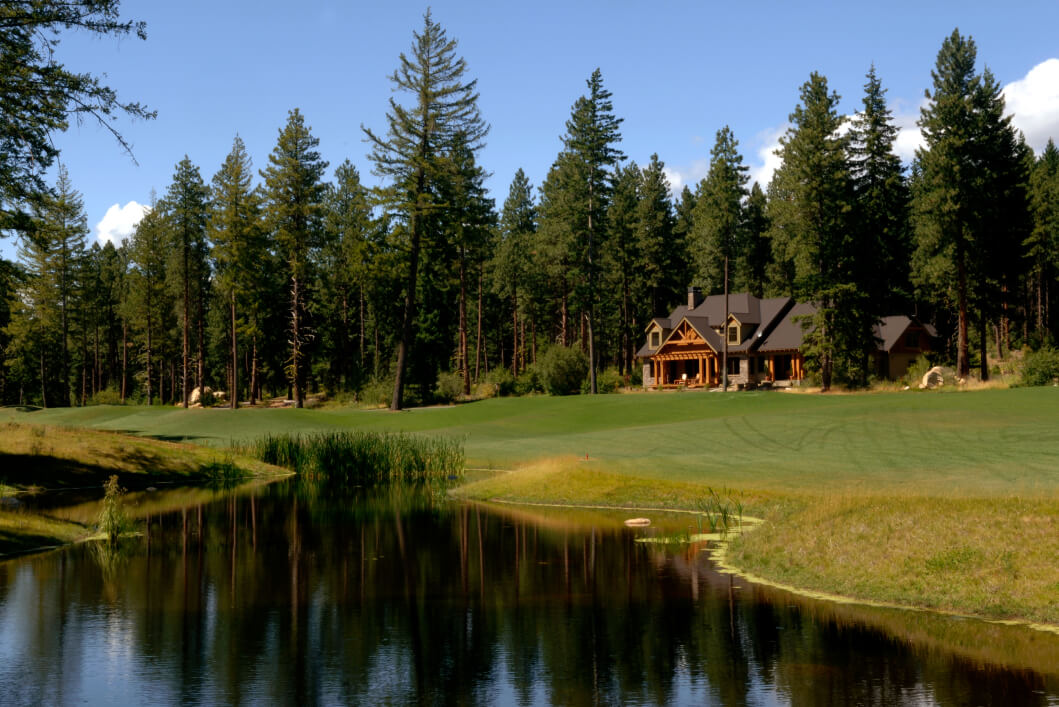 A large house with a wooden exterior is nestled among tall pine trees in Suncadia, set near a grassy lawn and a reflective pond under the clear blue sky.