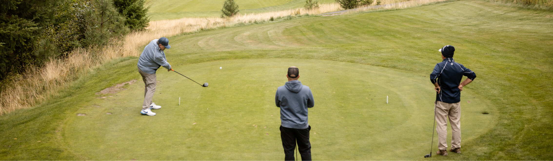 Three people enjoy a sunny day on the golf course in Suncadia. One is swinging a club at the tee, while the other two watch from behind, dreaming of nearby Suncadia homes surrounded by lush trees and grass.