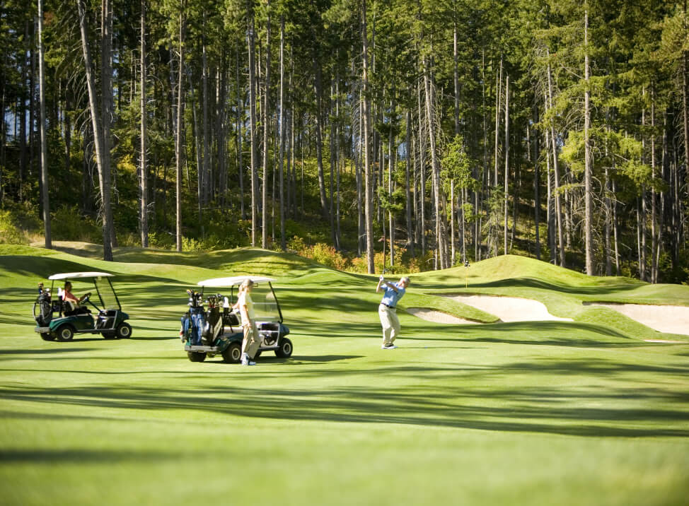 A person swings a golf club on a sunlit Suncadia golf course, where two golf carts await amid dense trees in the background. Nearby, Cle Elum restaurants tantalize with local flavors, adding to the allure of this picturesque setting.